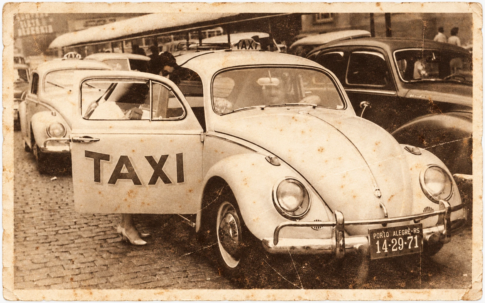 Fotografia de um táxi Volkswagen Fusca de porta aberta, diante de outros carros, em sépia, com superfície envelhecida e borda branca desgastada.